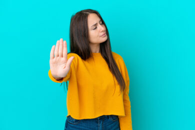 Image shows woman in yellow shirt on blue background with hand facing camera.