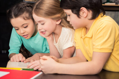 Image shows two girls and a boy lying on stomach reading together in a classroom.