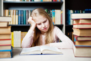 Image shows little girl with frustrated expression with an open book in front of her and stacks of books around her.