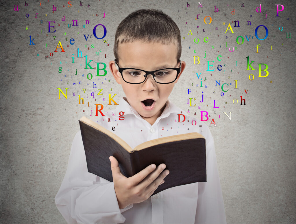 Image shows young boy in glasses and white shirt holding open a book with colorful letters floating off page.