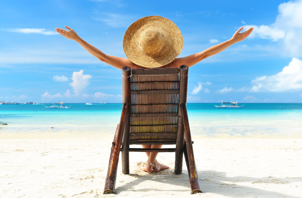 Image shows woman in straw hat sitting on brown chair on beach facing the water with her arms out, self-reflecting.