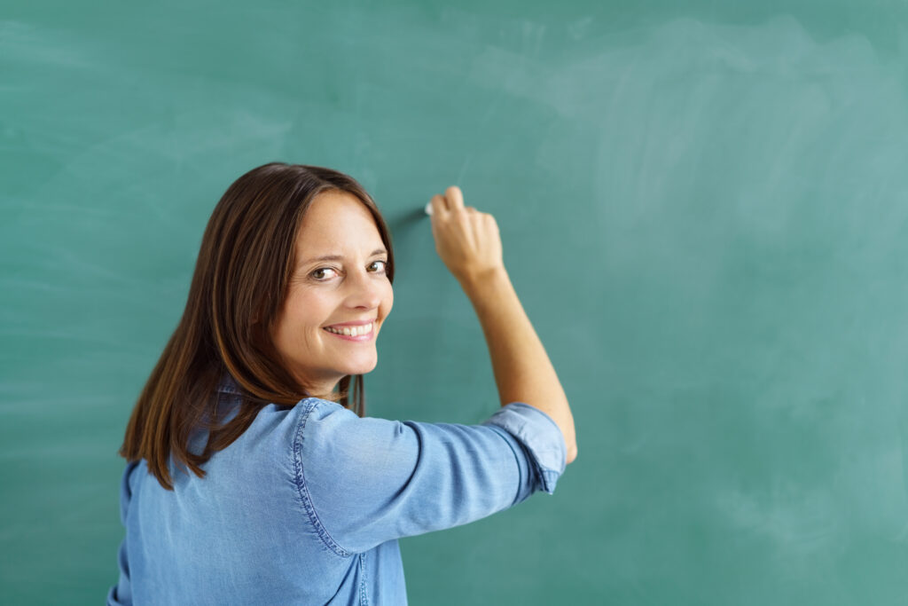 Teacher in blue shirt looking at camera and modeling writing on chalkboard.