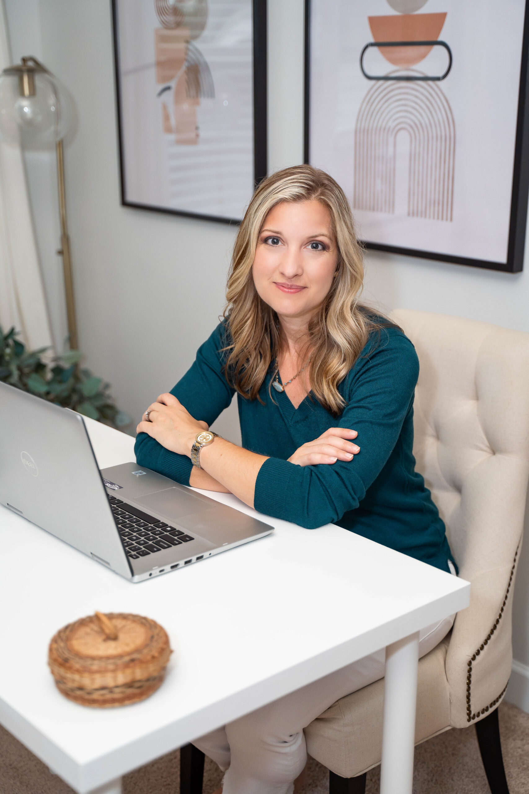 Blonde woman in teal sweater with arms folded in front of her resting on white desk with computer, showing that Coach from the Couch offers 1:1 virtual literacy support.
