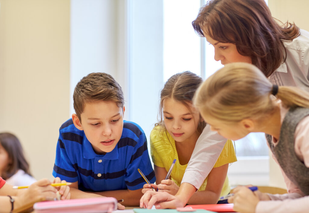 Teacher leaning over three students to  help with writing.