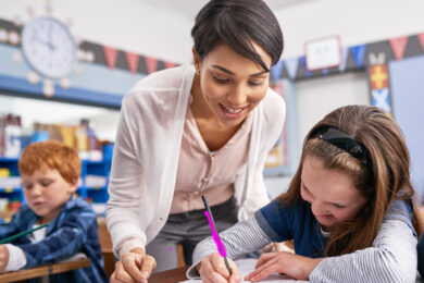 Teacher leaning over female student as she writes with pink pencil on white paper.