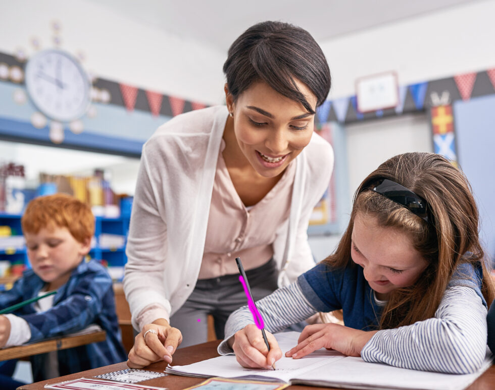 Teacher leaning over female student as she writes with pink pencil on white paper.