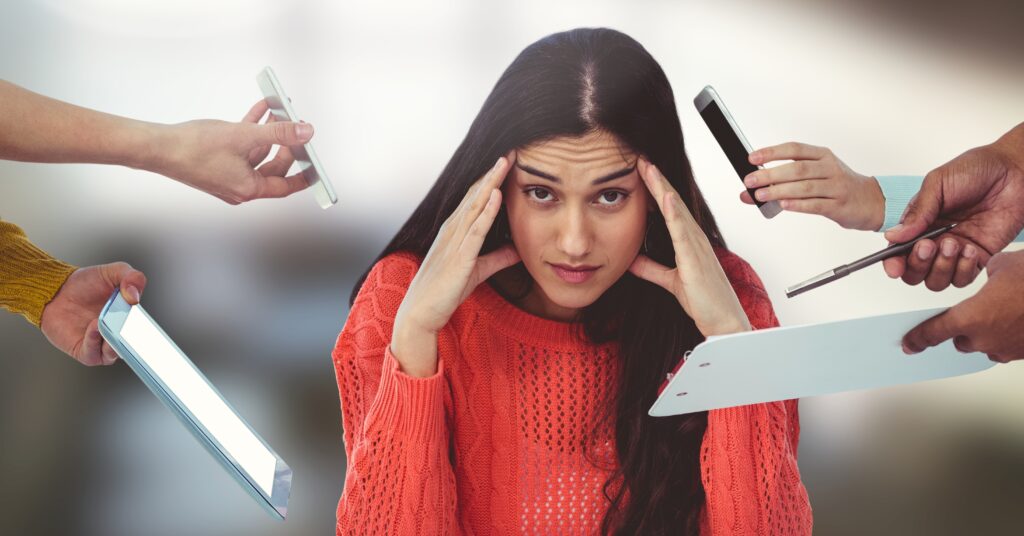 Image shows frustrated busy woman with brown hair in orange sweater with hands holding office supplies and devices reaching toward her.