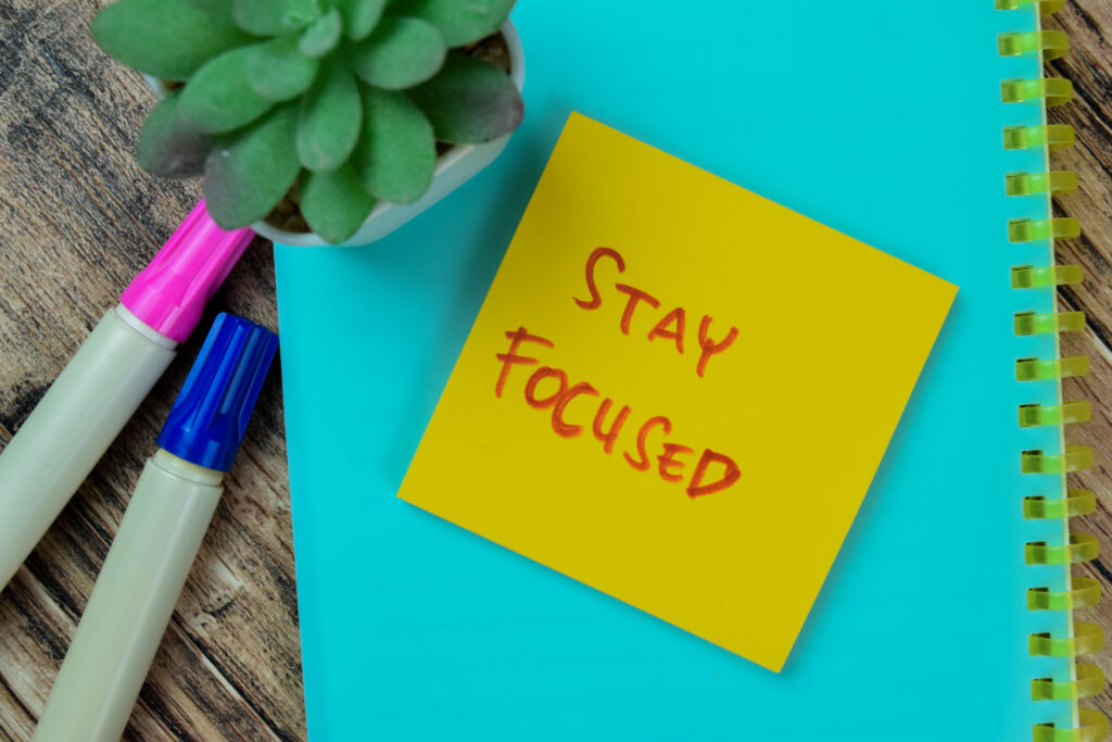 Image shows blue background of notebook with pink and blue marker and green plant on wood table with yellow post it note that says stay focused.
