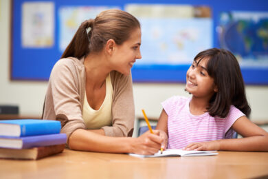 Image shows teacher with pencil in hand helping young student in pink shirt write.