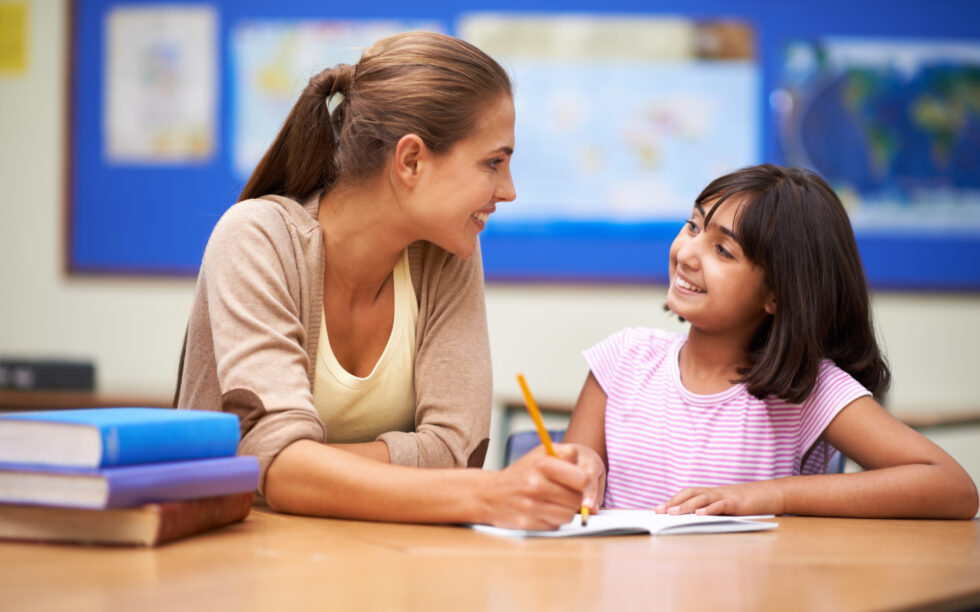 Image shows teacher with pencil in hand helping young student in pink shirt write.