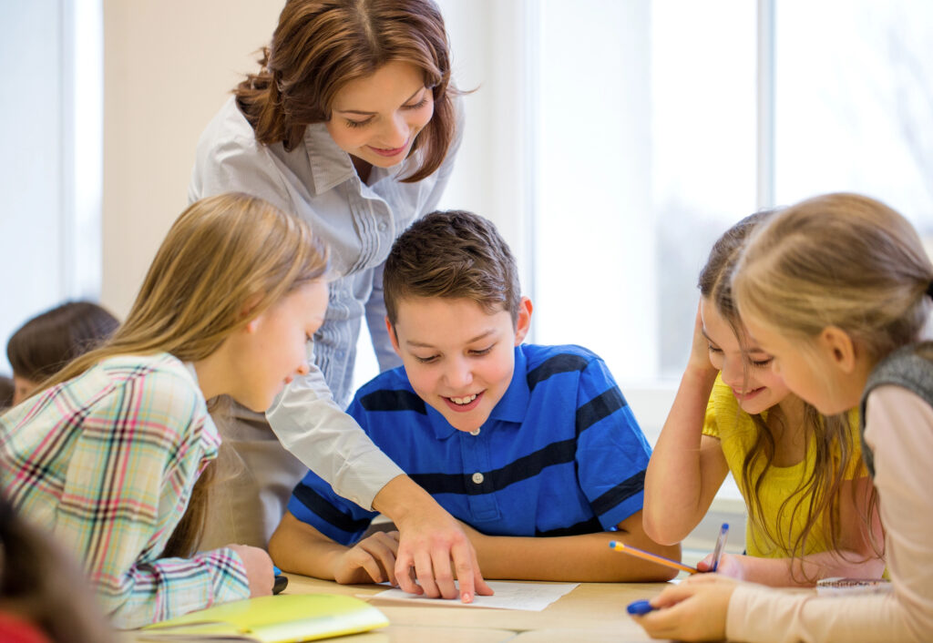 Image shows teacher standing behind group of students pointing to paper providing feedback to student about writing.