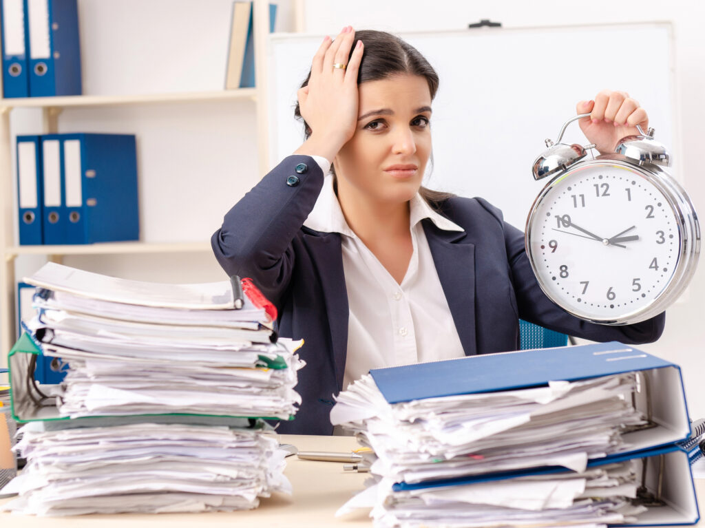 Teacher holding clock overwhelmed by stack of papers to grade. 