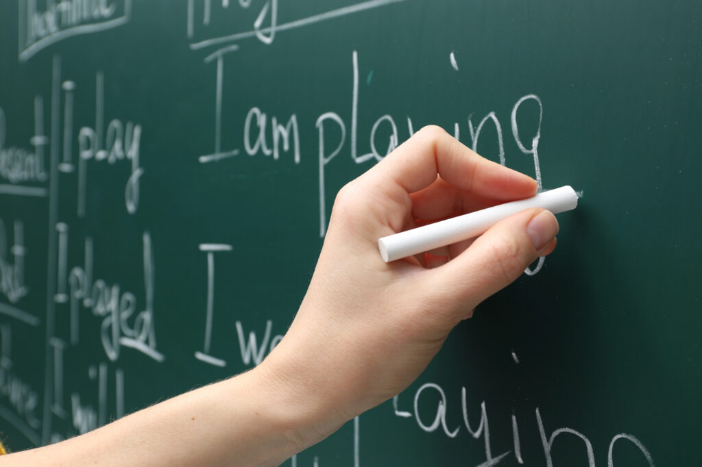 Hand holding chalk writing sentences that say I am playing to model sentence writing for students.