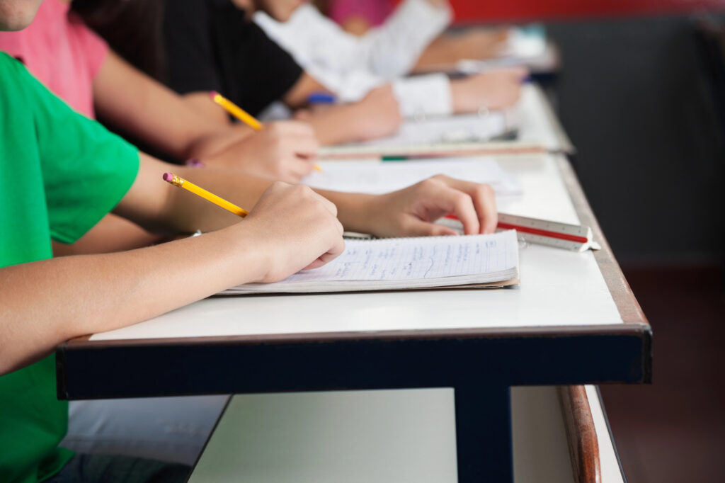Row of students in colorful shirts writing on notebook with a pencil at desks.  