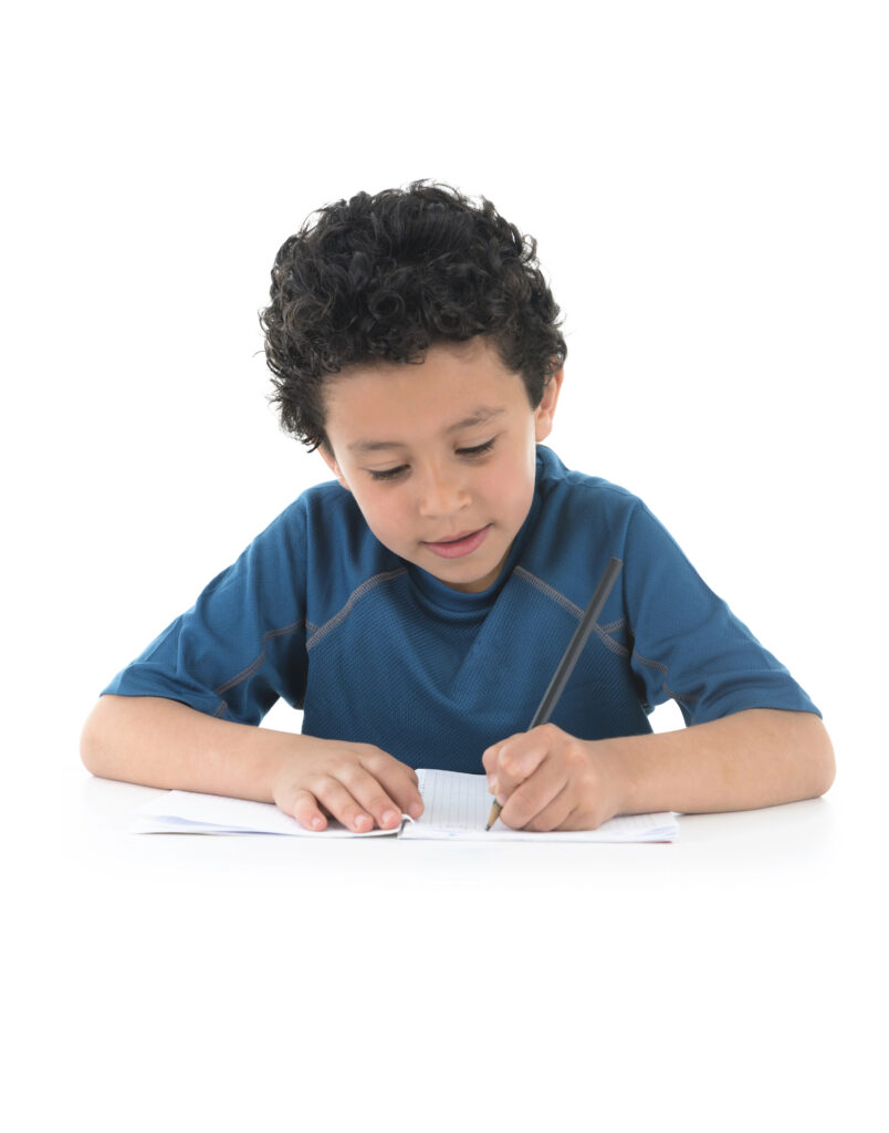 Young smiling Hispanic boy in blue shirt writing on white notebook with black pencil. 