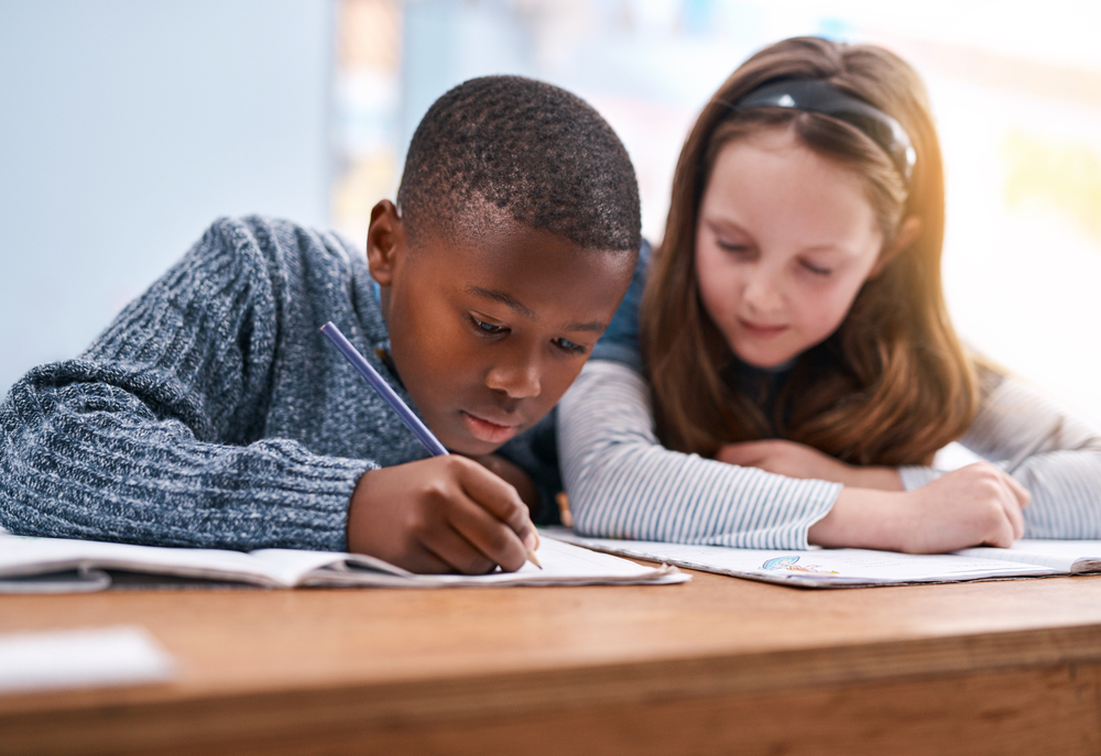 Black boy in blue sweater writing with blue pencil while caucasian girl in blue stripes helps.