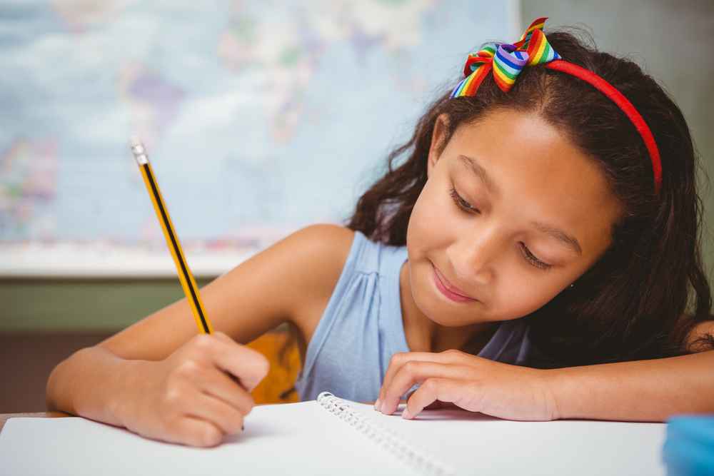 Hispanic girl in blue shirt with red headband writing with yellow pecnil on notebook with world map in background.