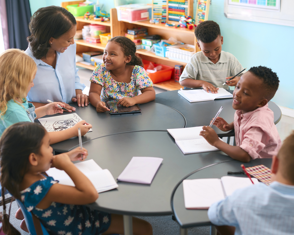 Black female teacher in blue shirt teaching vocabulary words at a table with multi-ethnic students.
