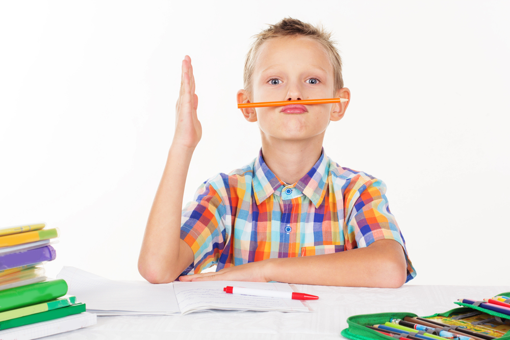 Boy in colorful shirt raising right hand and holding a pencil between nose and lips in front of school work.