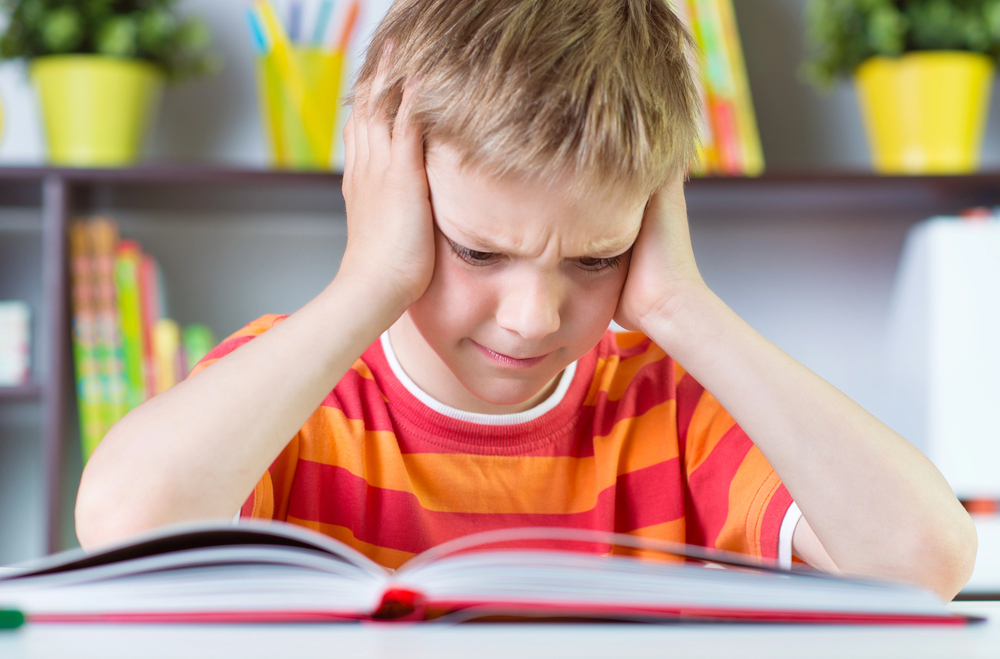 Image shows blonde boy in red and orange striped shirt with head in hands over a book frustrated to read out loud in class.