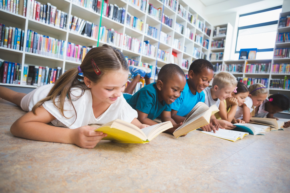 Row of multi-ethnic children lying on floor reading books and smiling in school library. 