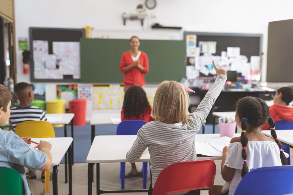 Female teacher in orange sweater in front of chalkboard facing classroom of students.