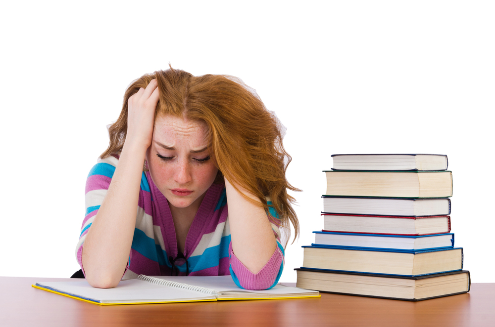 Girl with red hair and colorful striped sweater frustrated with head in hands over an open notebook next to a large stack of books.