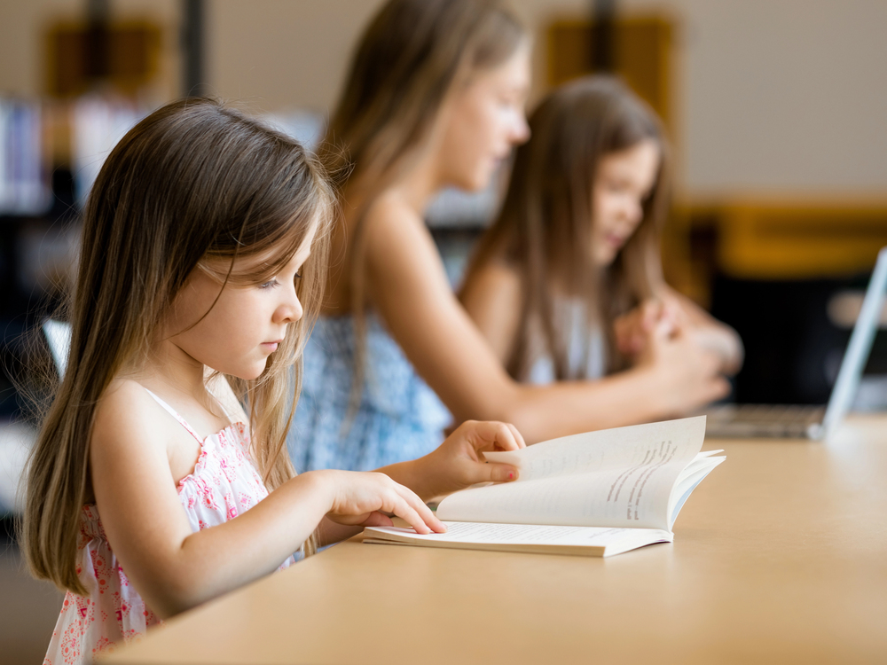 Young girl with brown hair and sleevless pink top reading book at table with second student and teacher on computer in background.