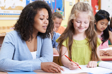 Image shows teacher with brown hair in blue sweater conferring about writing with female students with long blonde hair and green dress.
