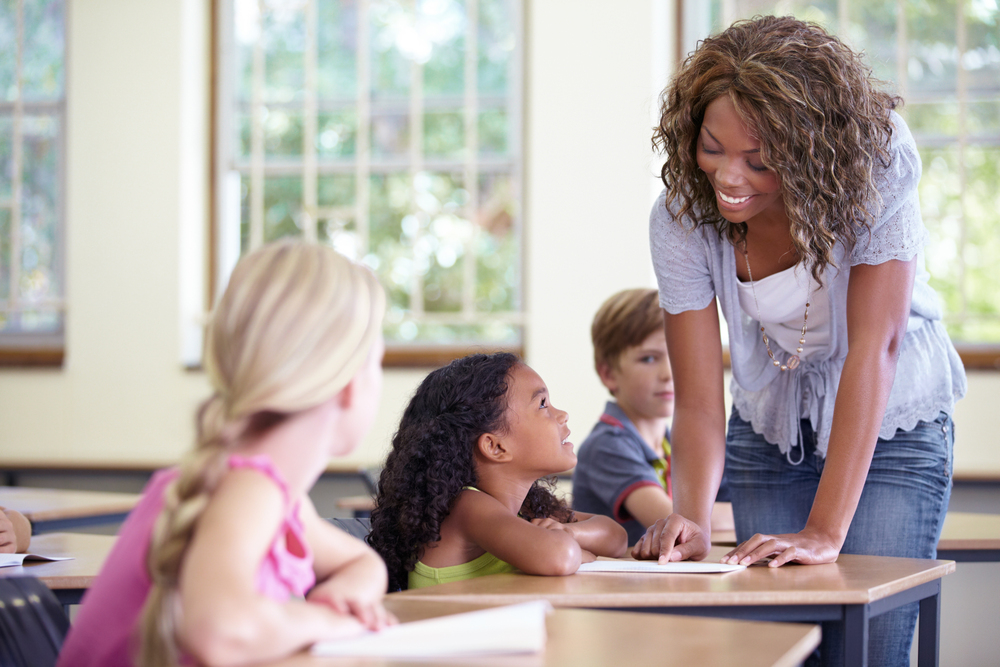Black teacher providing feedback to black female student to improve writing skills while two caucasian students listen in.