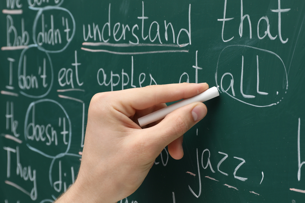 Caucasian teacher writing to model sentence construction with white chalk on green board. 