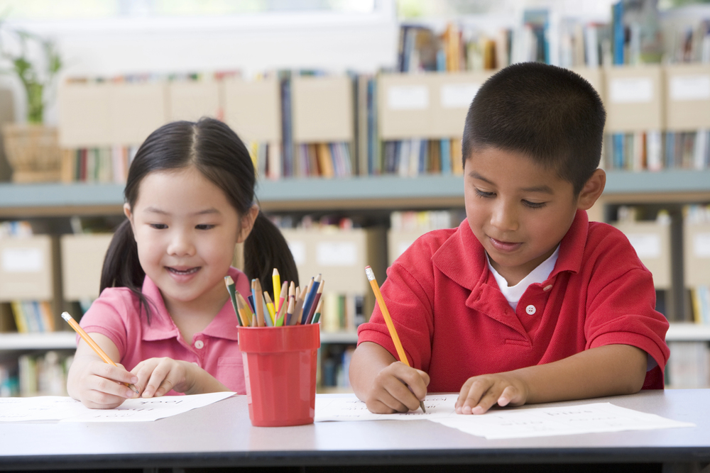Hispanic boy in red shirt and Asain girl in pink shirt next to red cup full of pencils writing about learning for purposeful review work.