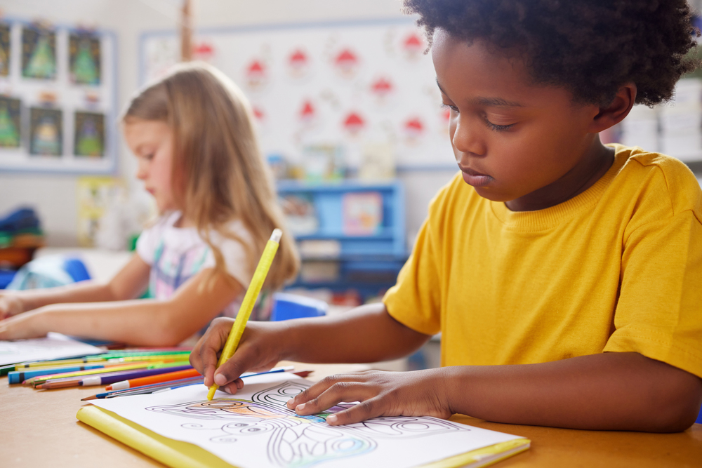 Black girl in yellow shirt and Caucasian girl in pastel dress miss out on learning by doing busywork of coloring a butterfly.  