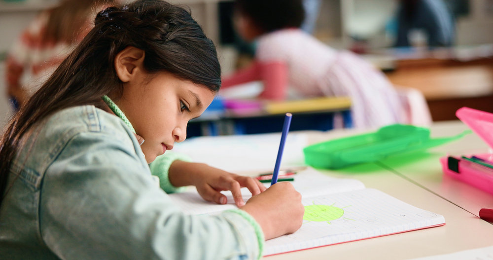 Young hispanic girl in denim jacket writing about previous learning with blue pencil on notebook paper in school.