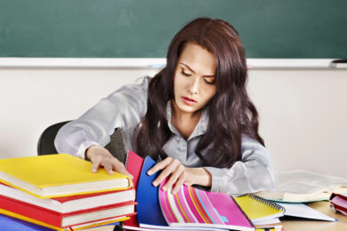 Image shows female teacher with brown hair and gray shirt looking through colorful student writing notebooks overwhelmed by teaching writing.