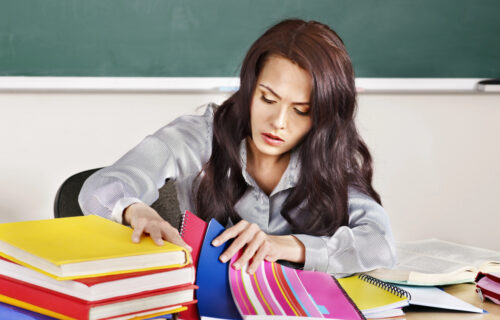 Image shows female teacher with brown hair and gray shirt looking through colorful student writing notebooks overwhelmed by teaching writing.