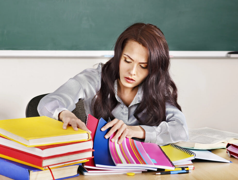 Image shows female teacher with brown hair and gray shirt looking through colorful student writing notebooks overwhelmed by teaching writing.