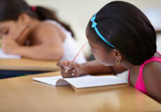 Multicultural female children engaged in writing in notebook with yellow pencil at school.