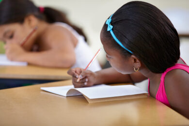 Multicultural female children engaged in writing in notebook with yellow pencil at school.