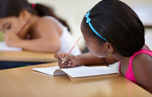 Multicultural female children engaged in writing in notebook with yellow pencil at school.