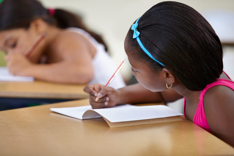 Multicultural female children engaged in writing in notebook with yellow pencil at school.