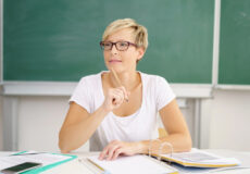 Female teacher wearing white shirt and classes tapping pencil on chin carefully planning data-driven reading lessons.
