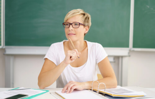Female teacher wearing white shirt and classes tapping pencil on chin carefully planning data-driven reading lessons.