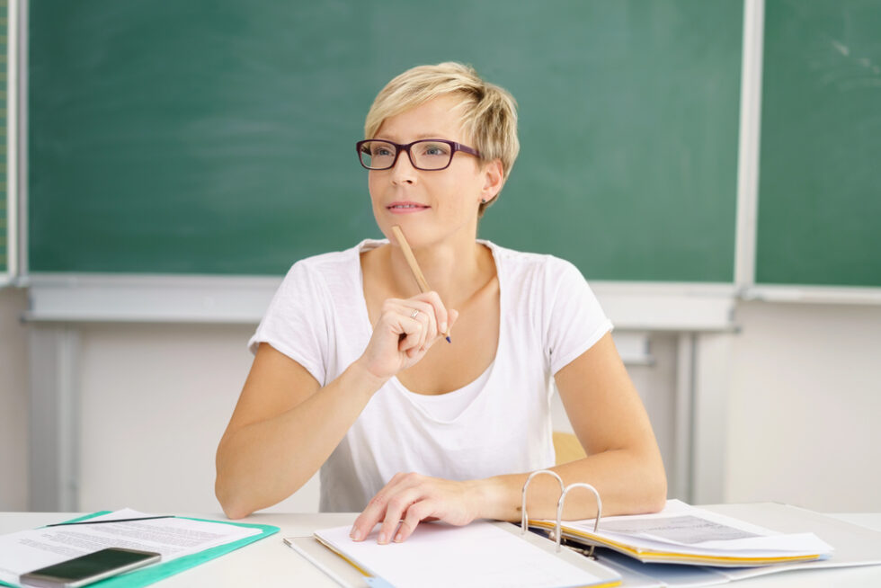 Female teacher wearing white shirt and classes tapping pencil on chin carefully planning data-driven reading lessons.