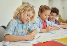 Schoolchildren wearing blue writing sentences with pencils in notebooks at school.