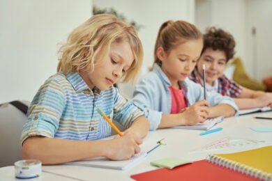 Schoolchildren wearing blue writing sentences with pencils in notebooks at school.